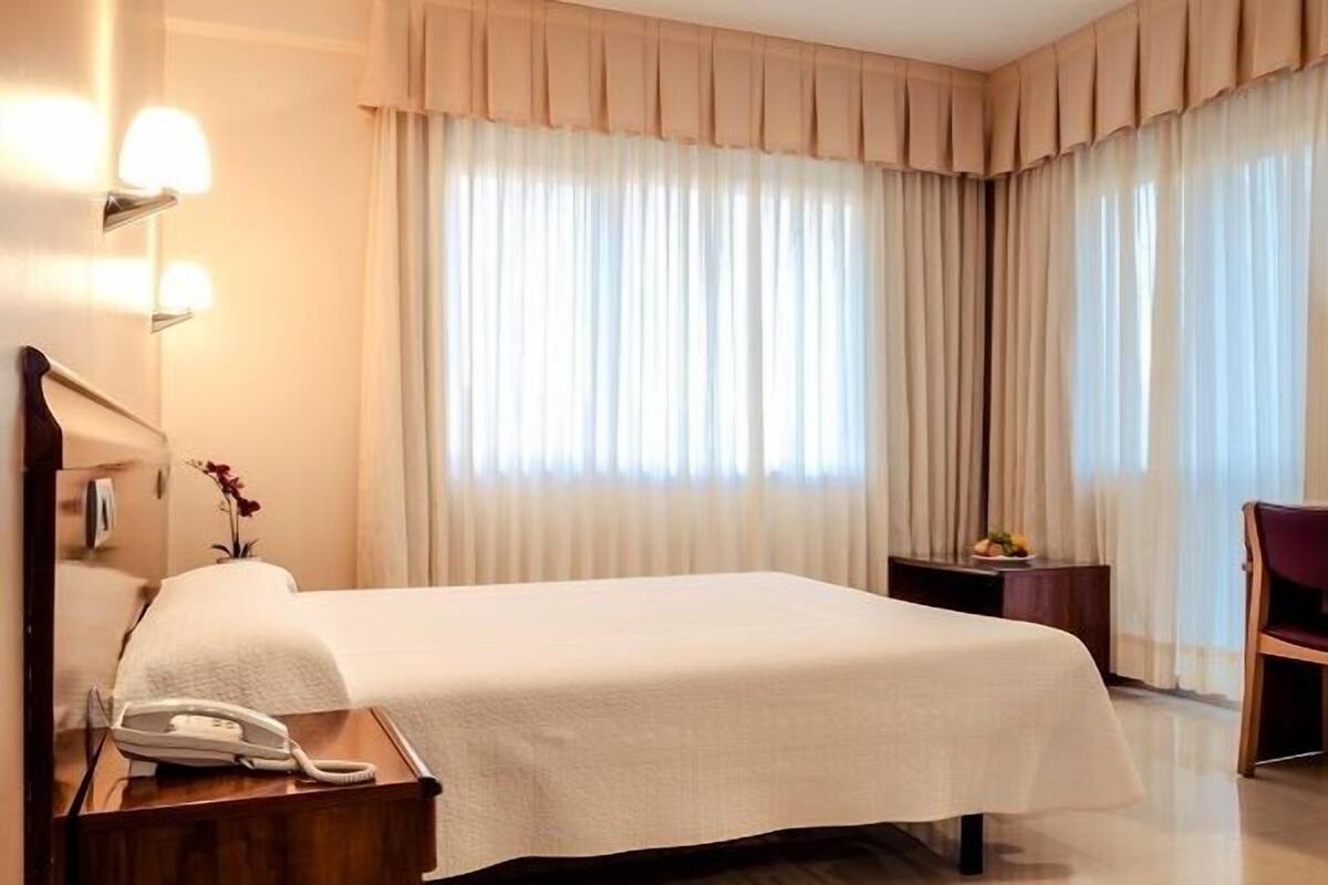 Een hotelkamer met een bed, nachtkastje met een telefoon, een klein tafeltje met een vaas en fruit, en een stoel.