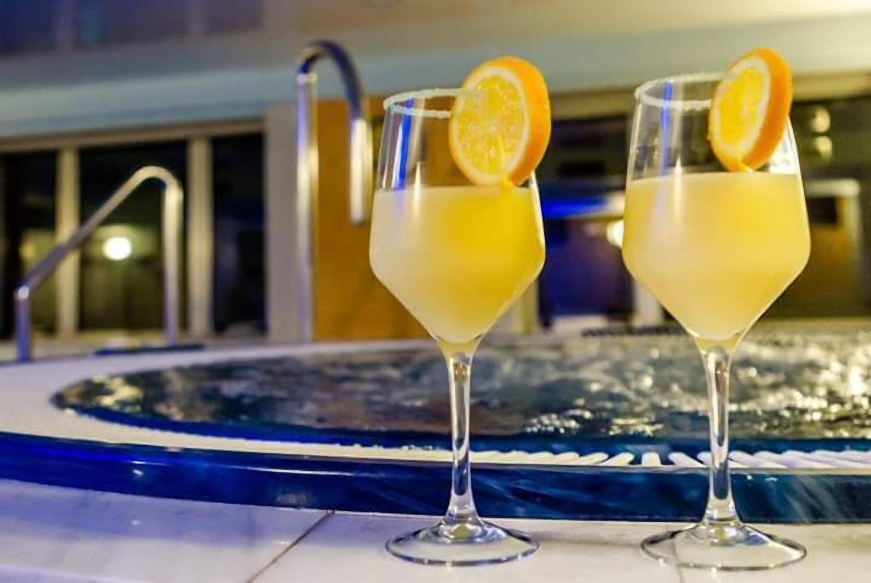 Dos cócteles con rodajas de naranja sobre una superficie reflectante cerca de una piscina.