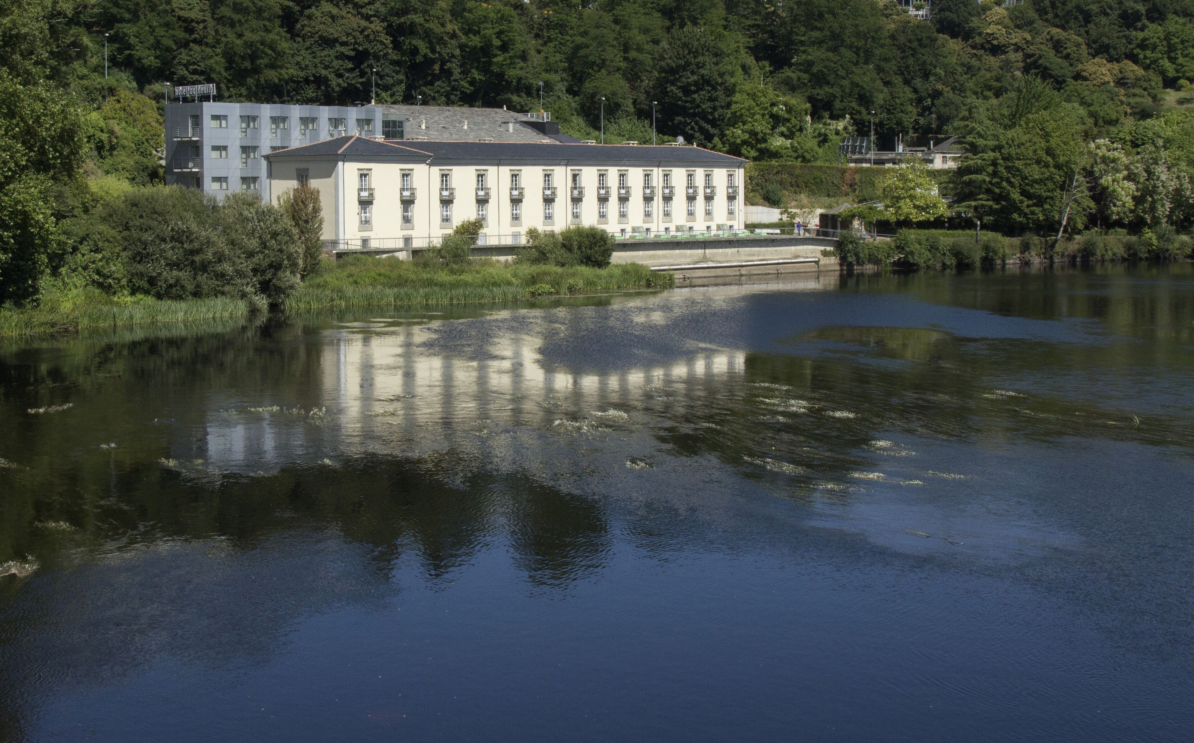Un edificio grande con múltiples ventanas y balcones, ubicado junto a un cuerpo de agua con reflejos.