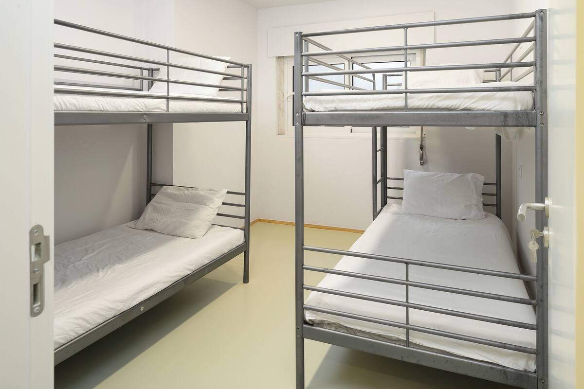 Dos literas con barandillas metálicas y ropa de cama blanca en una habitación con pared lisa y una ventana.