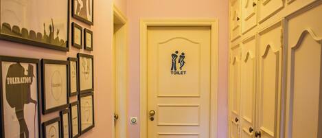 Een gang met ingelijste kunstwerken aan de wanden, een deur met het symbool voor een toilet en een rij kastjes aan de rechterkant.