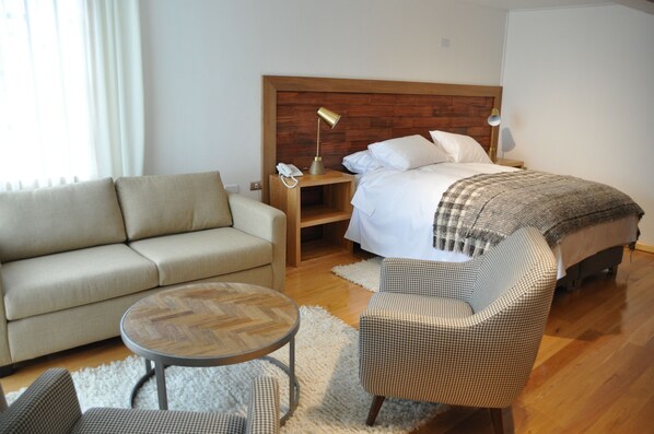 A hotel room with a wooden headboard bed, a beige sofa, a round coffee table, and a checkered armchair.