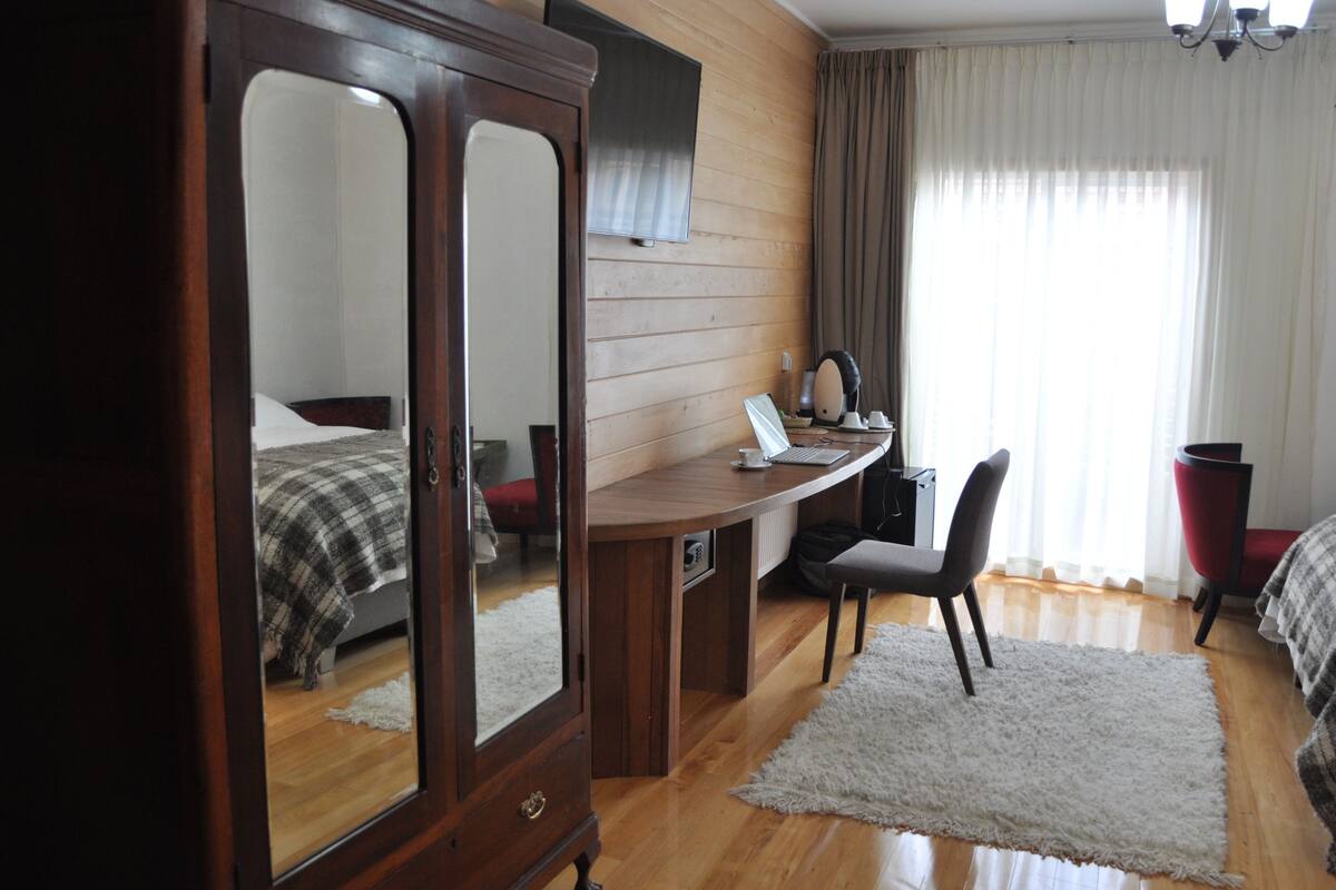 A room with a wooden wardrobe, a desk with a chair, a bed with a checkered blanket, and a large window with curtains.