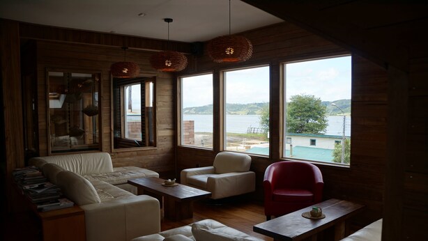 A cozy cabin-style room with a large window offering a view of a lake and hills. There are several white leather sofas and a red chair. - Hotel Boutique Refugio de Navegantes (Dalcahue)