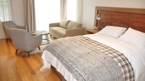 A hotel room with a large bed, a wooden headboard, a grey armchair, a beige sofa, a small round table, and a lamp on a wooden nightstand. - Hotel Boutique Refugio de Navegantes (Dalcahue)