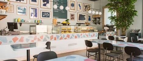 A cafe with a counter displaying food items, framed artwork on the walls, and multiple seating areas with tables and chairs.