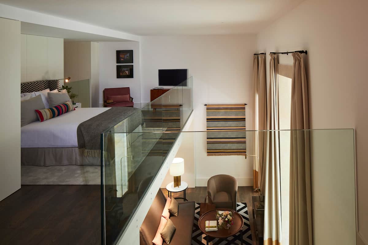 Una camera d'hotel moderna con un balcone in vetro, un letto con molti cuscini, una TV e un'area salotto con un tavolo e una sedia.