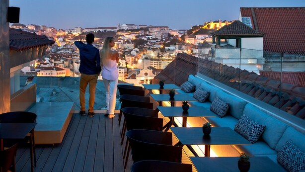 A couple standing on a rooftop terrace overlooking a cityscape at dusk. The terrace features a sofa, multiple tables, and chairs.
