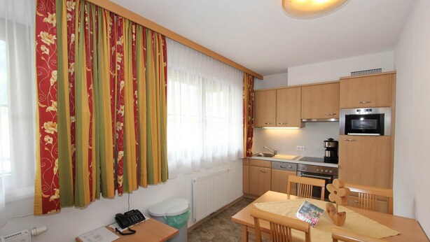 A compact kitchen with a stove, oven, and a coffee maker. There is a dining table with chairs and a small window with curtains.