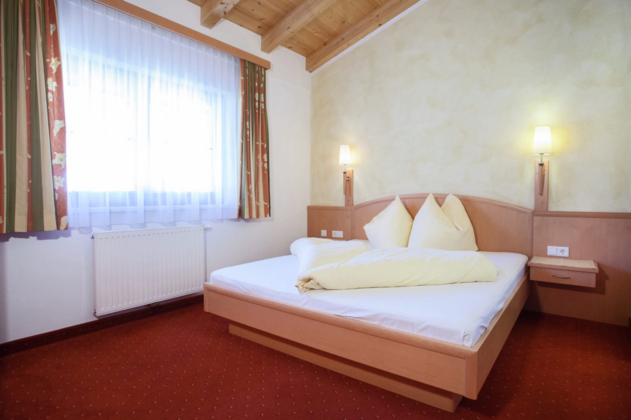A hotel room with a bed, two bedside lamps, a window with curtains, and a radiator.
