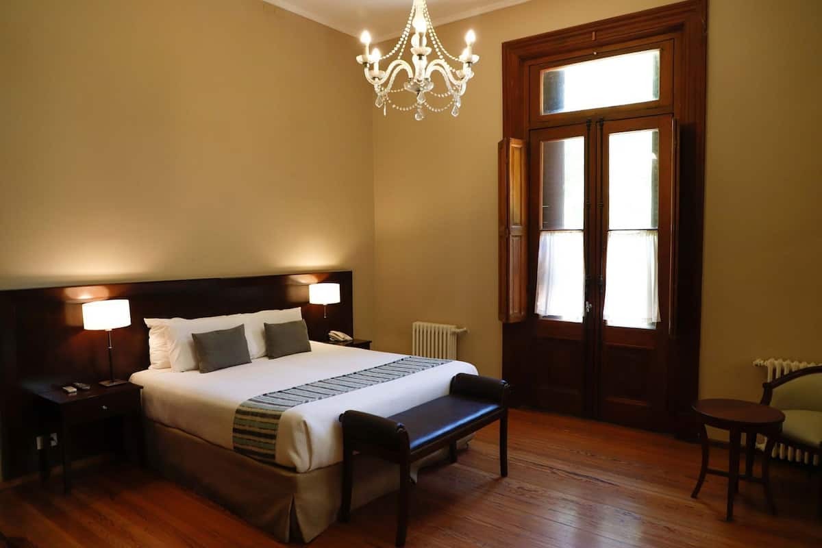 A hotel room with a large bed, bedside lamps, a wooden bench, a chandelier, a window with curtains, and a radiator.