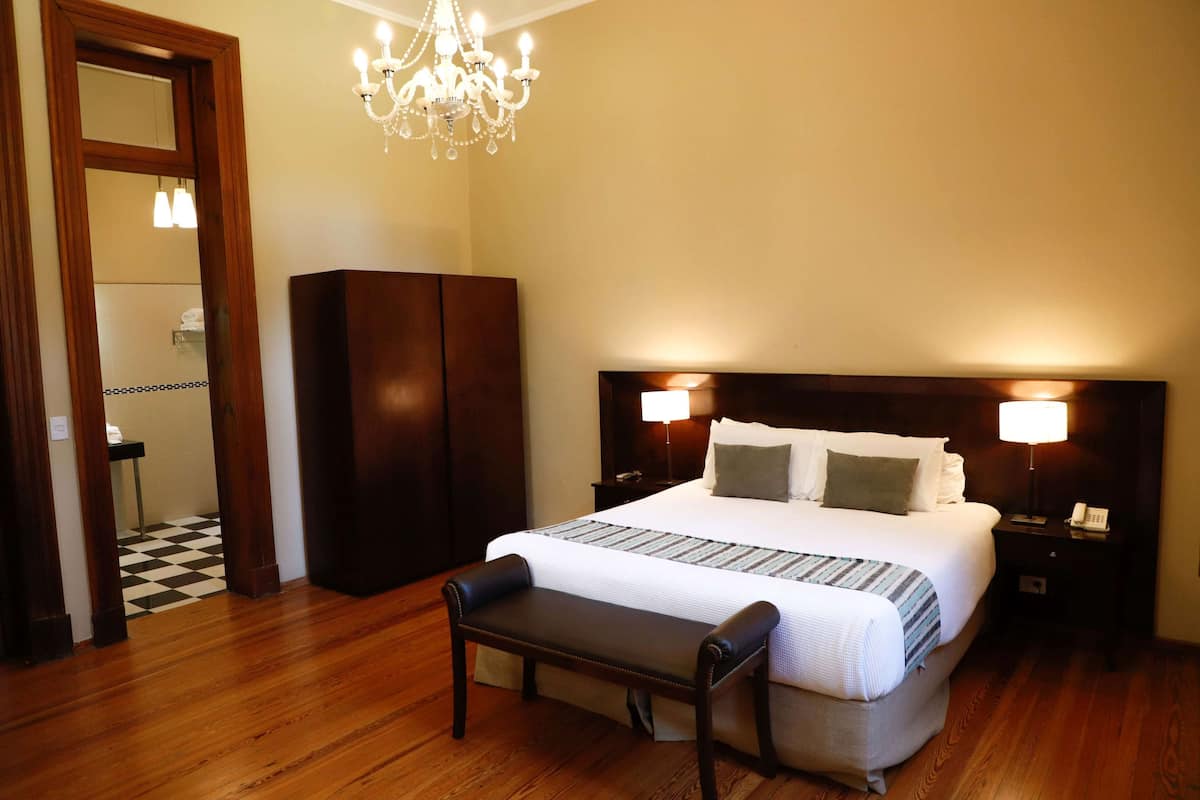 A hotel room with a large bed, wooden floor, a bench, a chandelier, and a nightstand with a lamp.