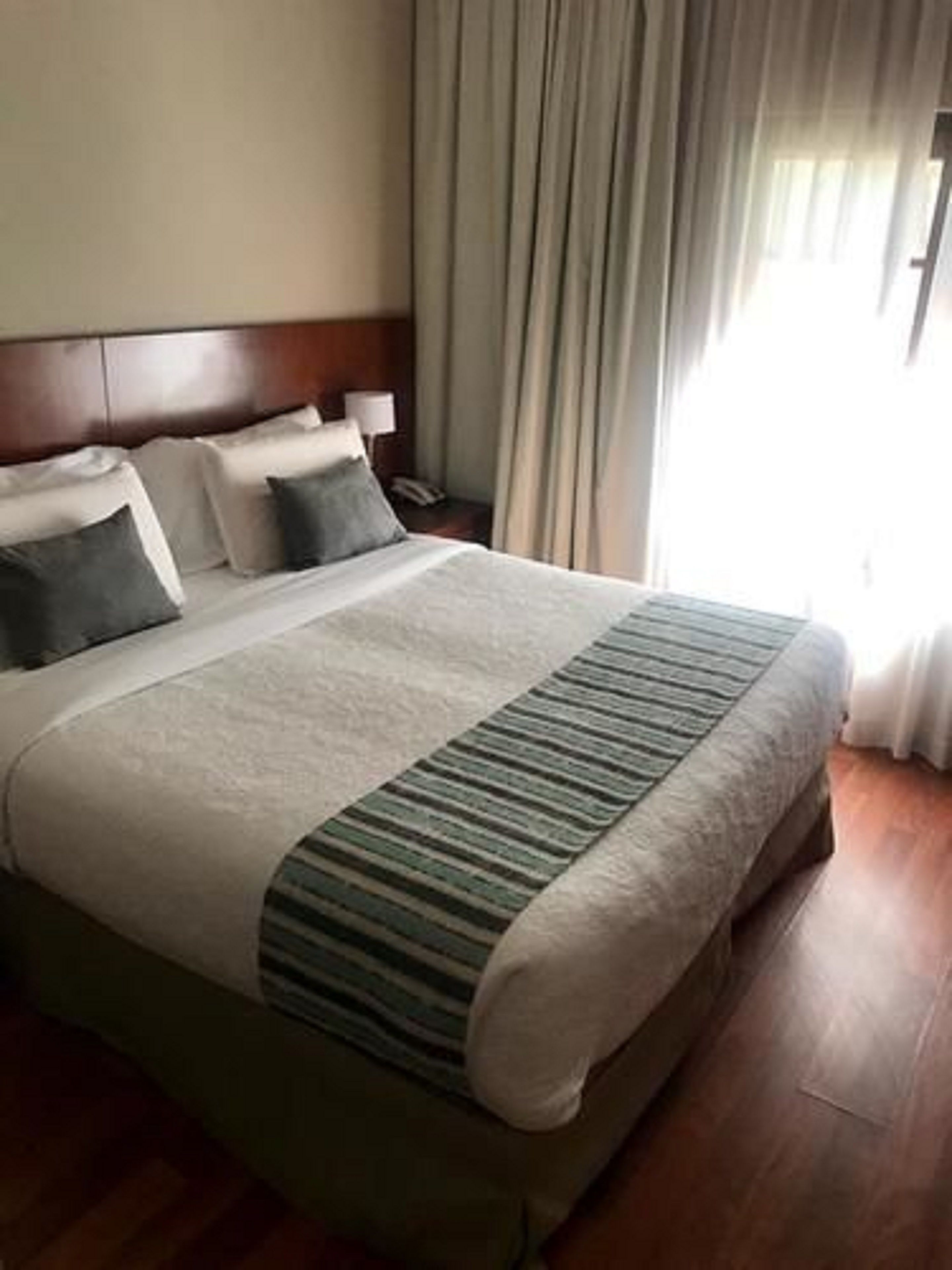 A hotel room with a large bed, a wooden headboard, a nightstand, and a window with curtains.