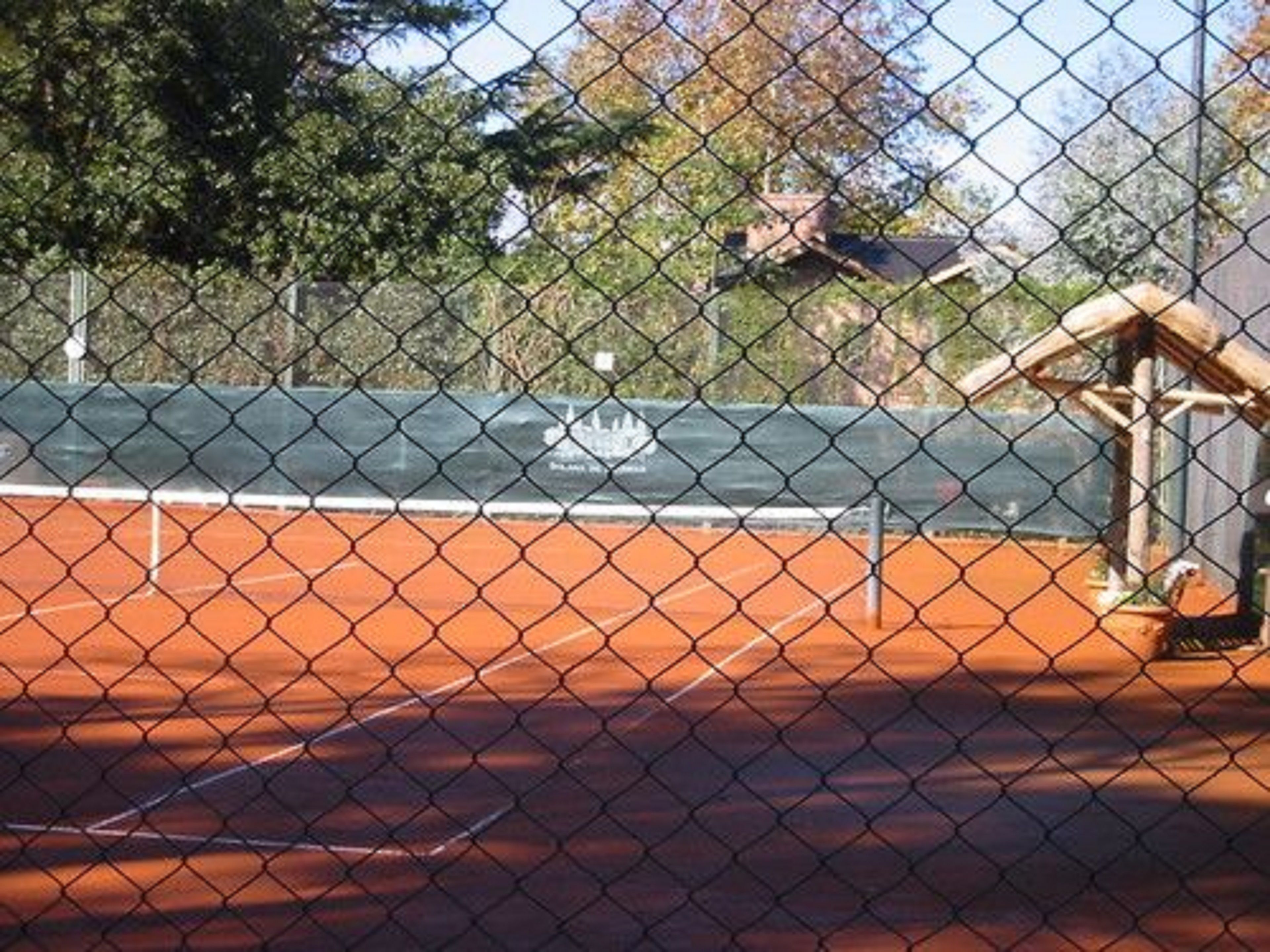 A tennis court with a chain-link fence, surrounded by trees and a building structure.