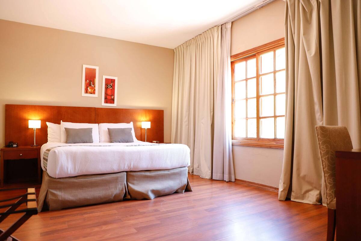 A hotel room with a large bed, two bedside lamps, a wooden headboard, a window with curtains, and a nightstand with a lamp.