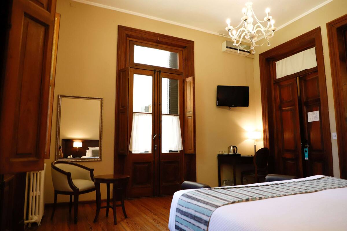 A hotel room with a bed, a chair, a small table, a TV, and a chandelier.
