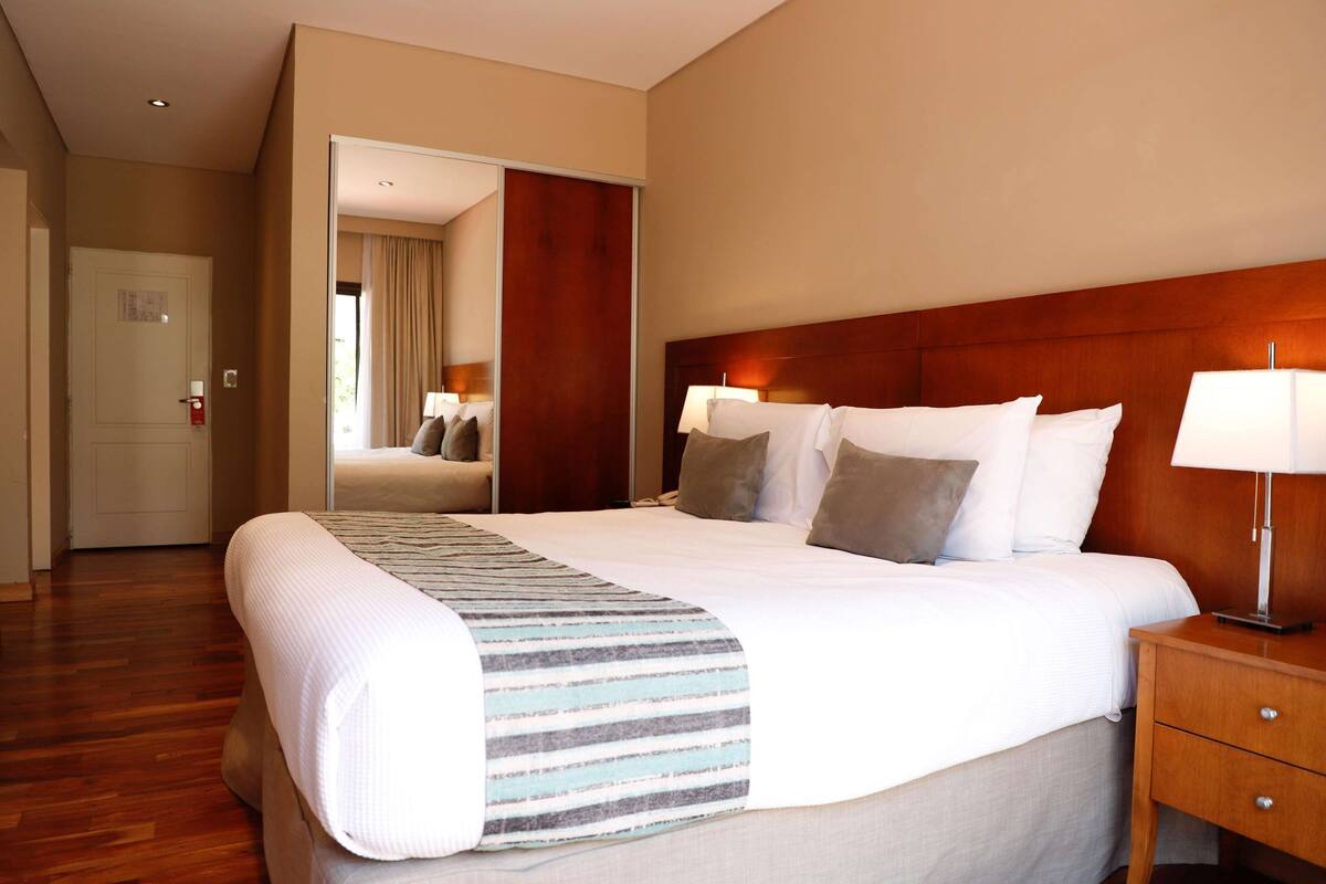 A hotel room with a large bed, wooden headboard, two bedside lamps, a wooden nightstand, and a door leading to another room.
