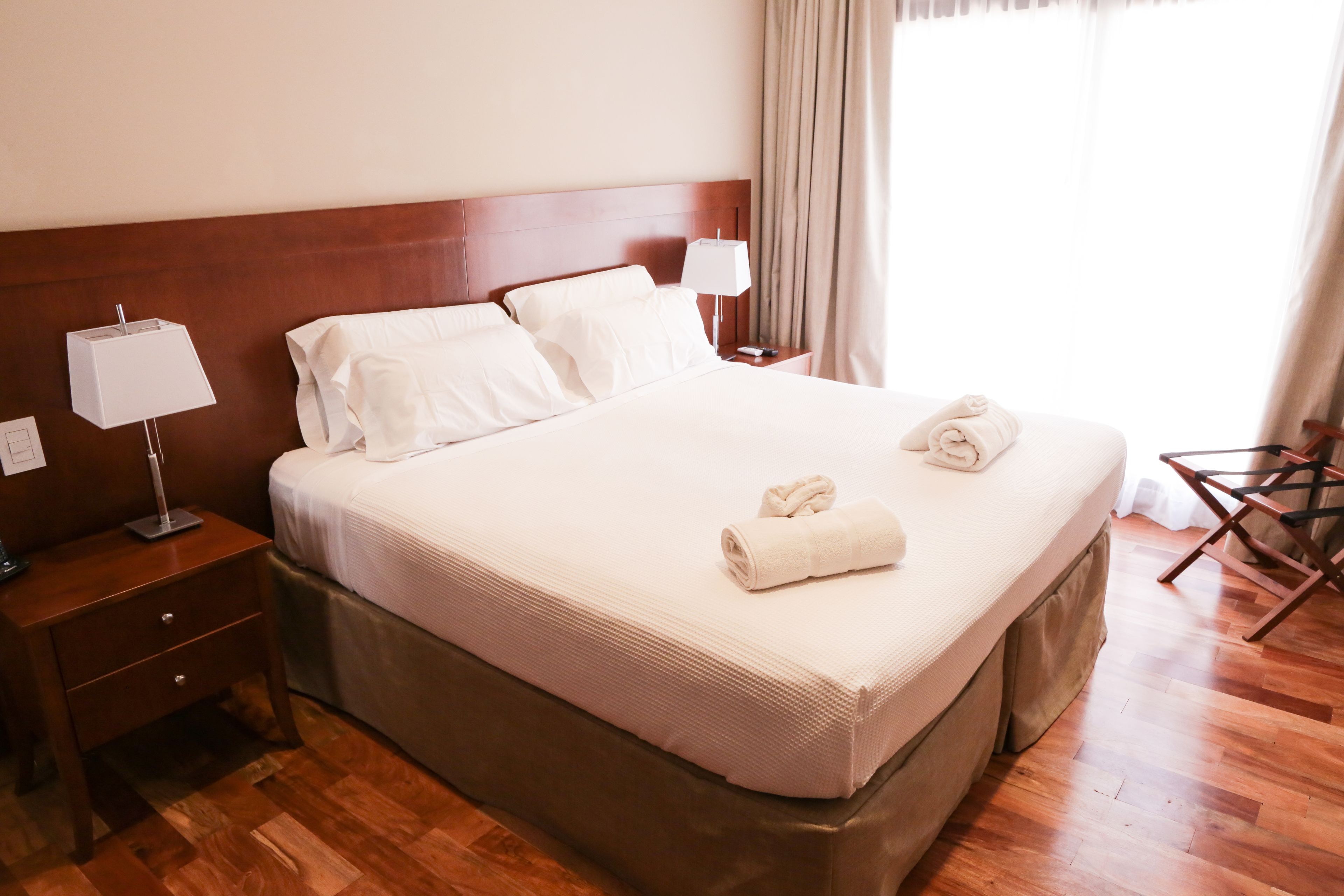A neatly made bed with white linens and two rolled towels, a wooden nightstand with a lamp, and a wooden chair.