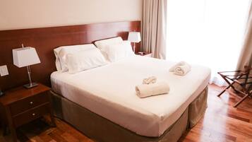 A neatly made bed with white linens and two rolled towels, a wooden nightstand with a lamp, and a wooden chair.