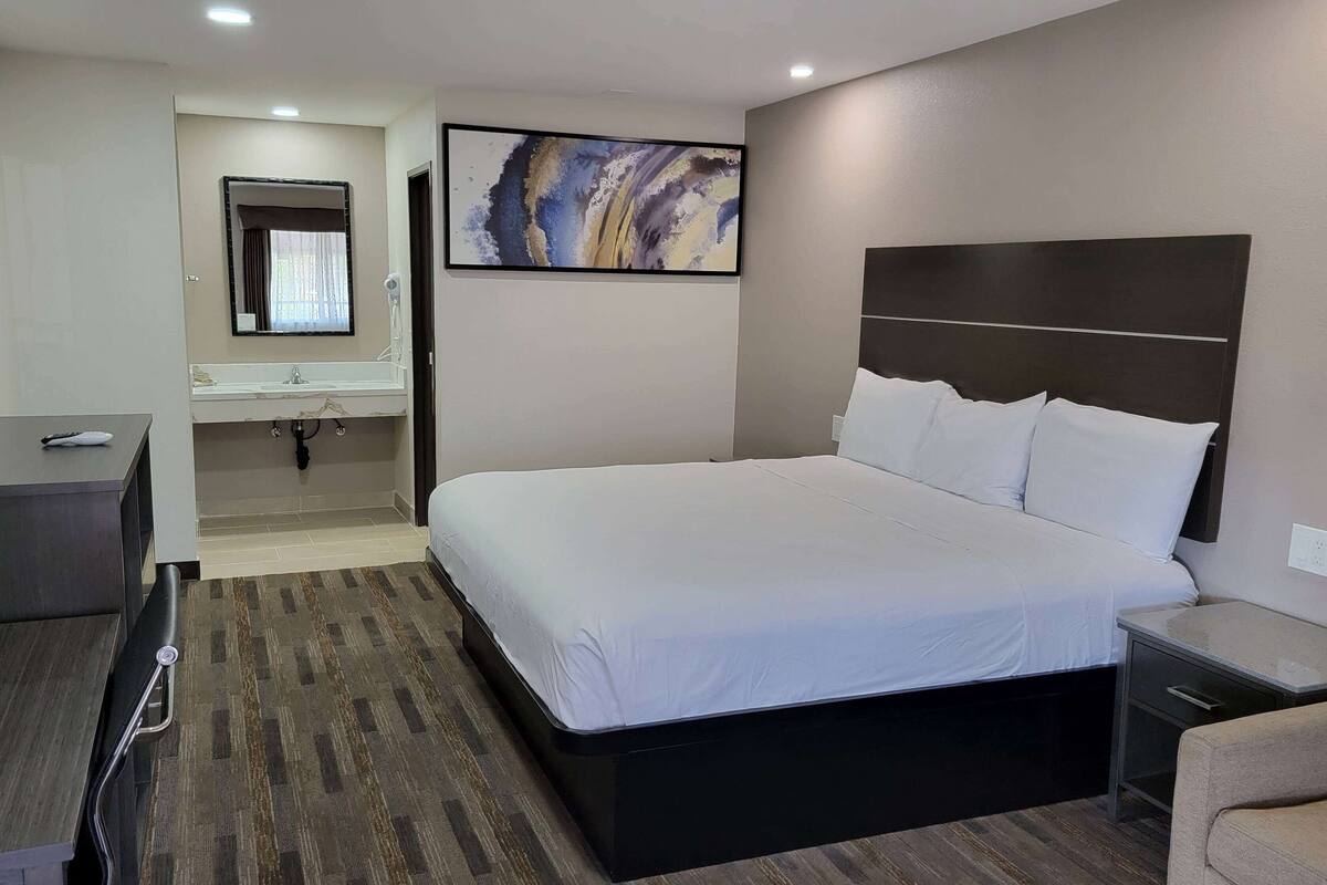 A hotel room with a large bed, a bedside table, a desk, a chair, a bathroom with a mirror, and a painting on the wall.