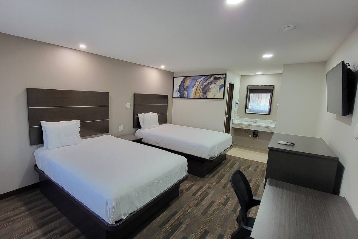 A hotel room with two beds, a desk, a chair, a TV, and a bathroom with a mirror and sink.