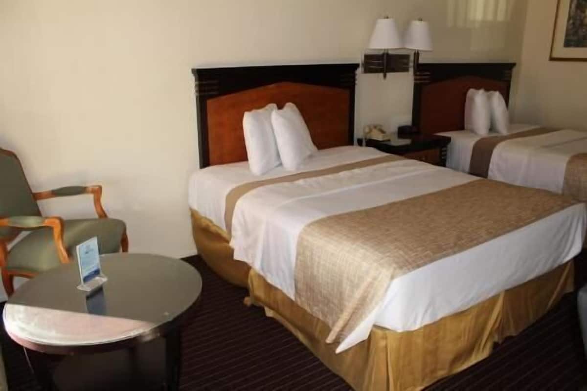 A hotel room with two beds, a chair, a small table, and a lamp.