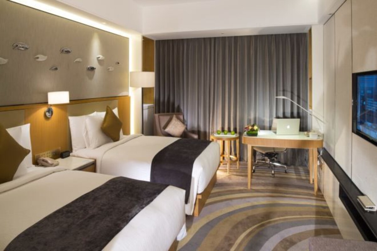 A hotel room with two beds, a desk with a laptop, a TV, and a large window with curtains.