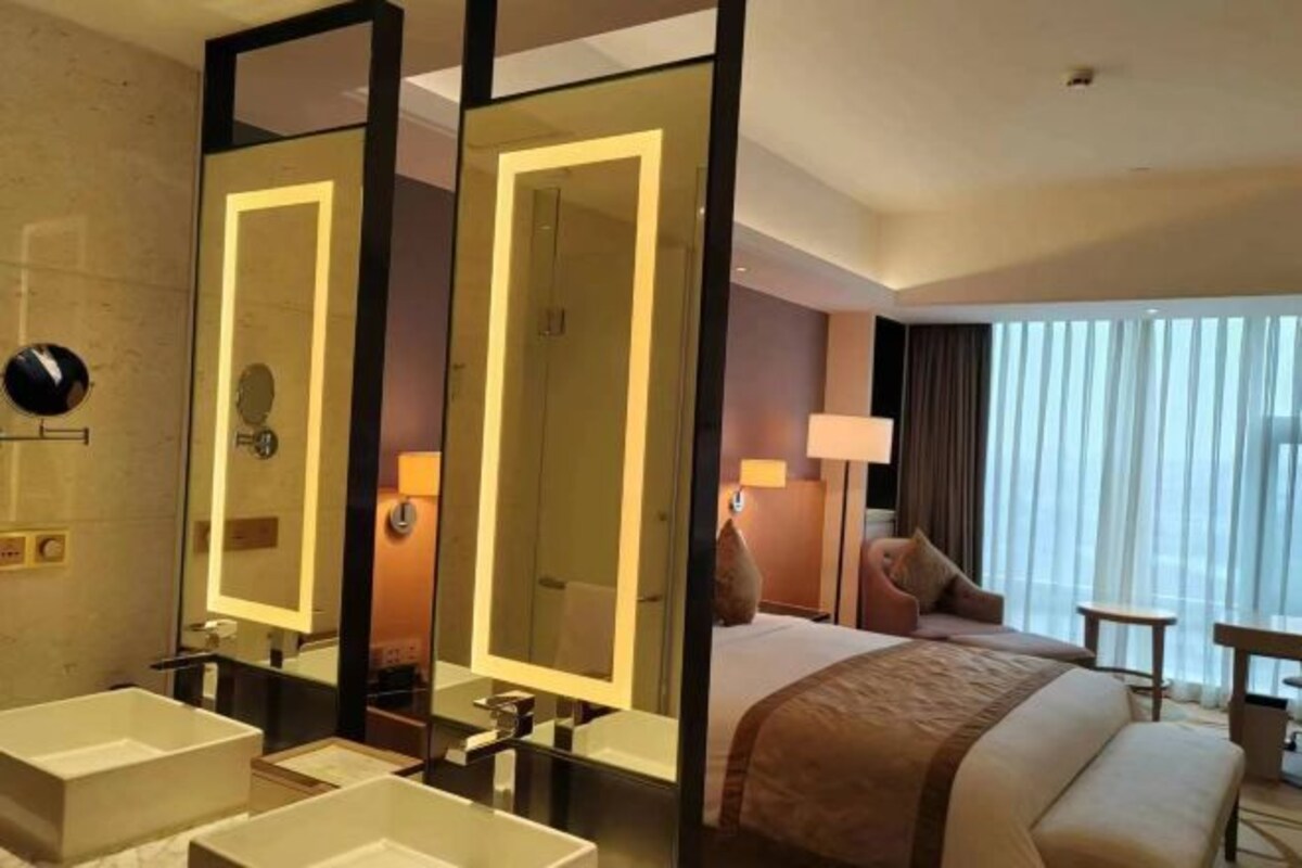 A hotel room with a large bed, two bedside tables, a desk, and a chair. There are two sinks with mirrors and a large mirror above the bed.