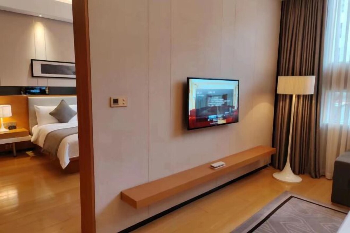 A hotel room with a bed, a TV mounted on the wall, a bedside table with a lamp, and a wooden bench.