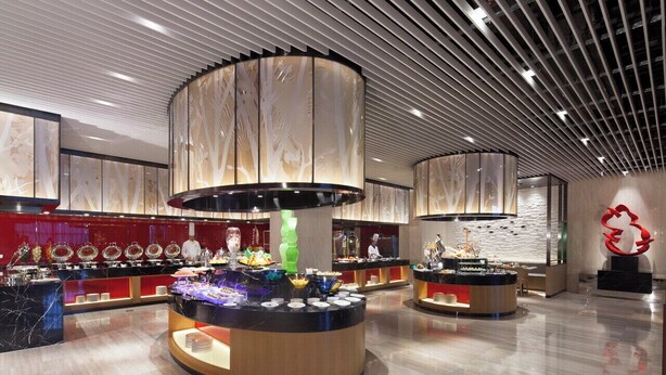 A modern hotel lobby with a central buffet area, a large abstract sculpture, and a polished marble floor.