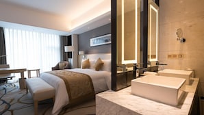 Een moderne hotelkamer met een groot bed, een zithoek, een bureau, een grote spiegel en een badkamer met bad en wastafel.