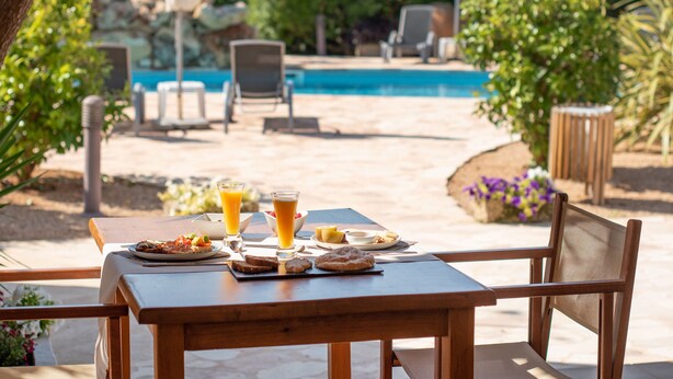 A wooden table with breakfast items and drinks, a pool in the background, and outdoor seating.