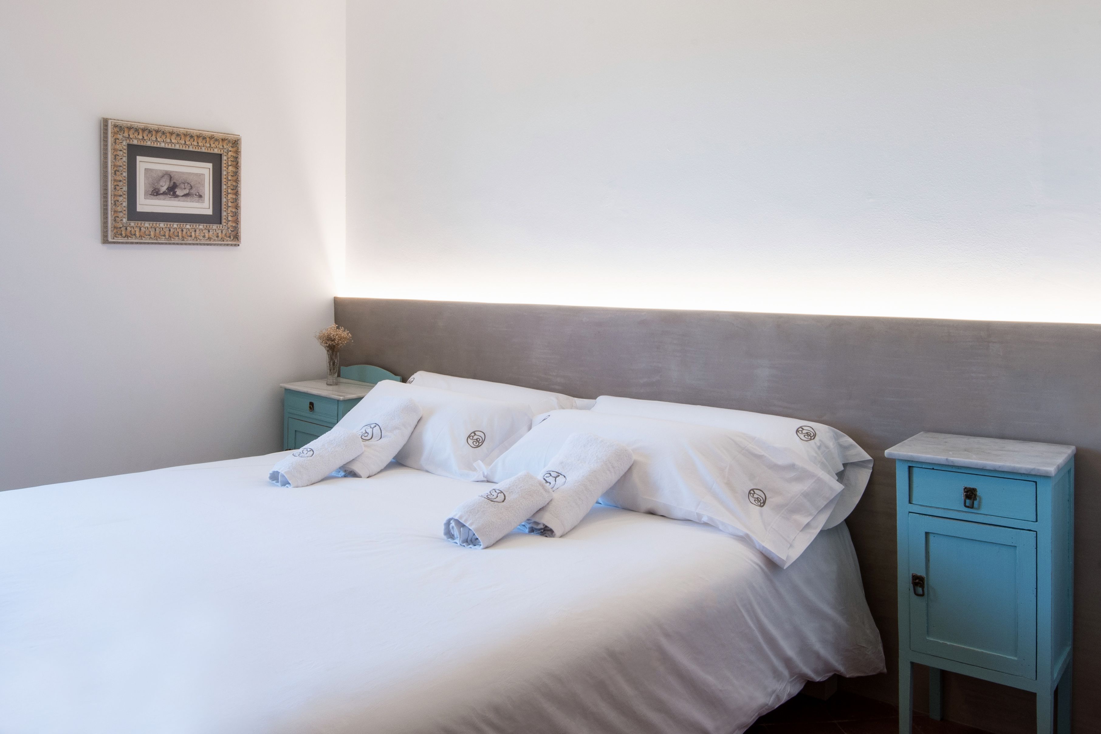 Una cama bien hecha con sábanas y toallas blancas, una mesita de noche con cajón y un cuadro en la pared.