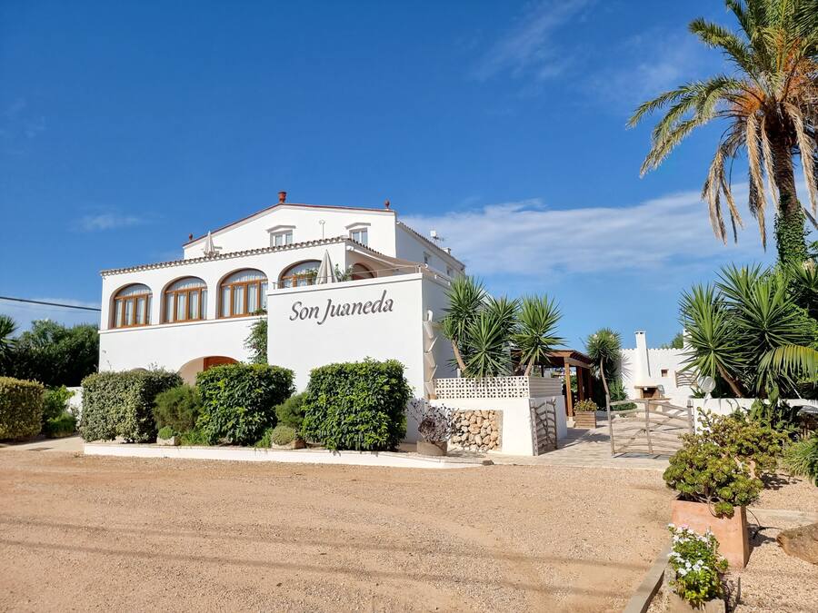 A white building with the name "Son Juaneda" on the facade, surrounded by greenery and palm trees.