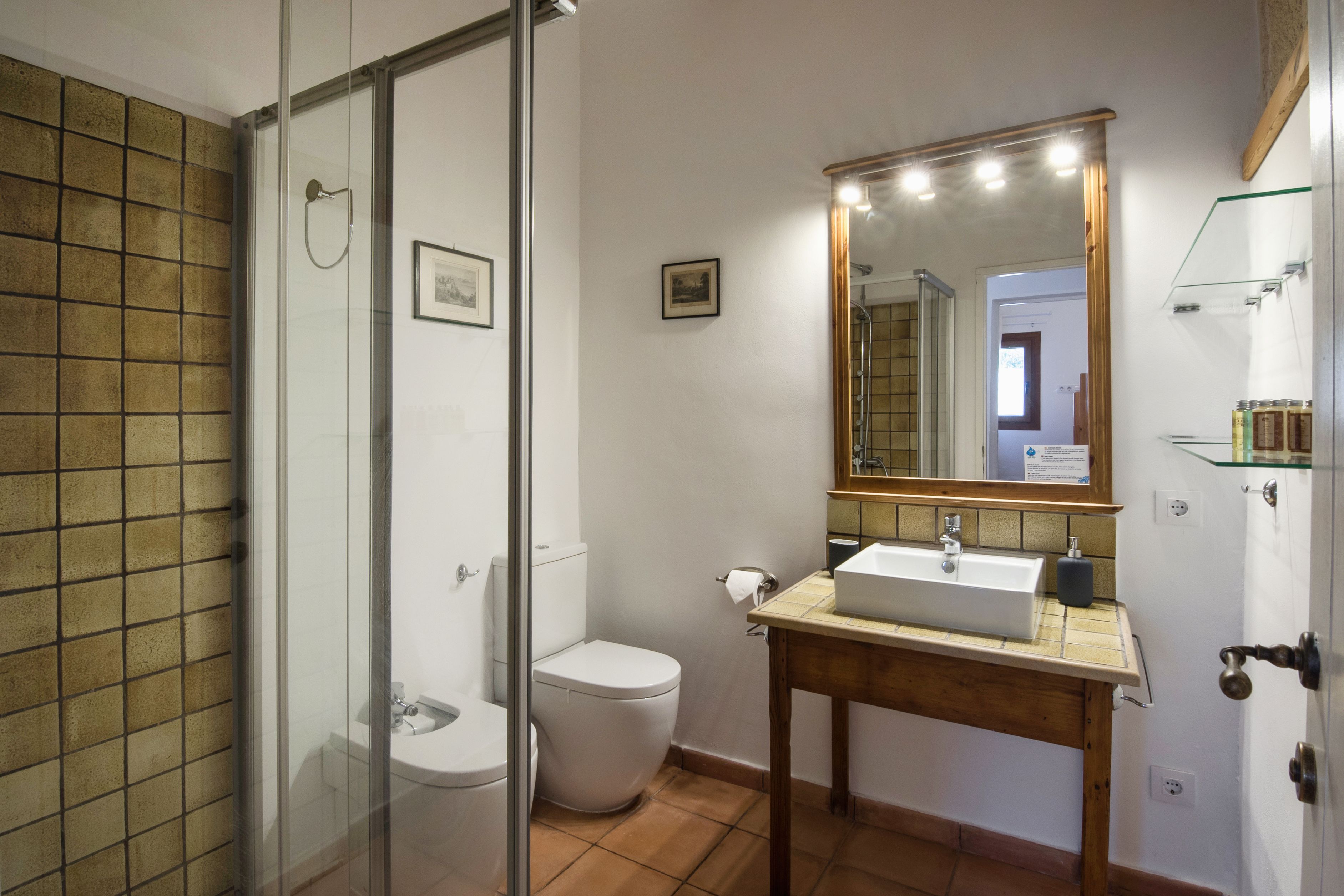 A bathroom with a glass shower enclosure, a white toilet, a wooden vanity with a sink, and a mirror with lights.
