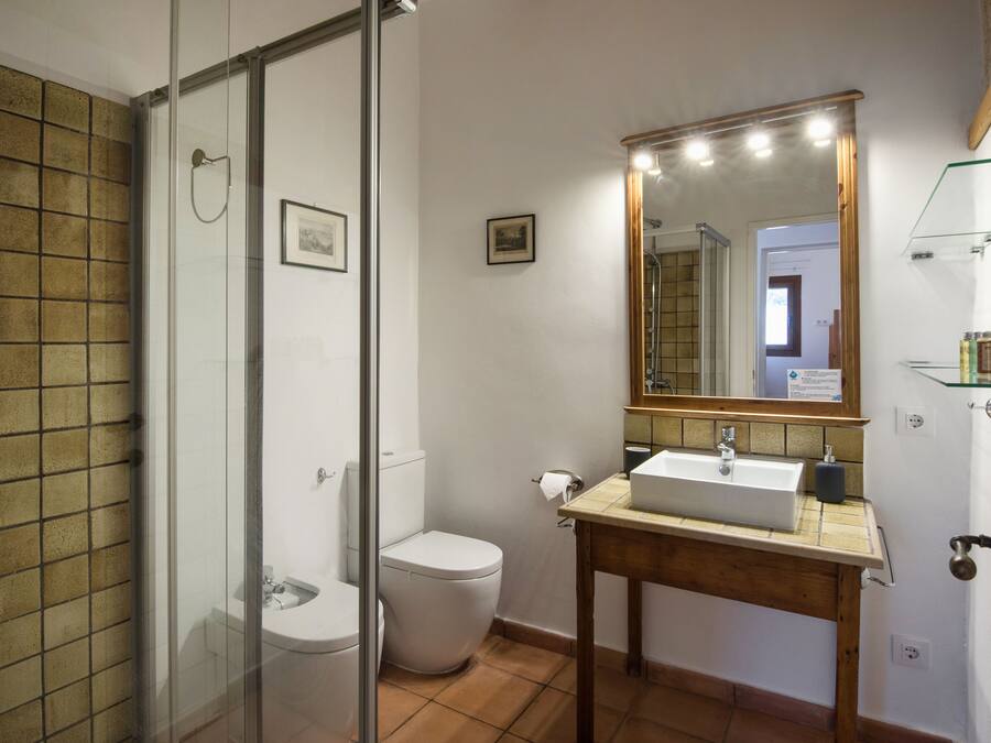 A bathroom with a glass shower enclosure, a white toilet, a wooden vanity with a sink, and a mirror with lights.