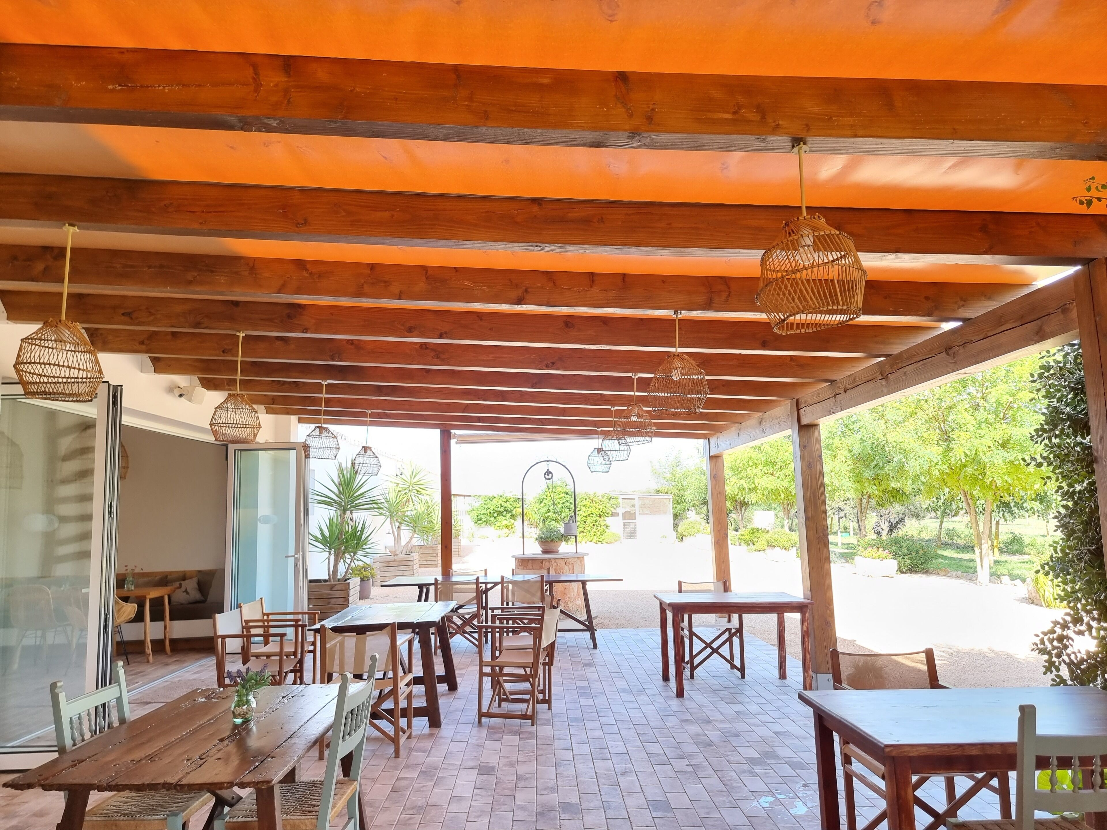 An outdoor patio with wooden tables and chairs, hanging wicker lamps, and a view of greenery.