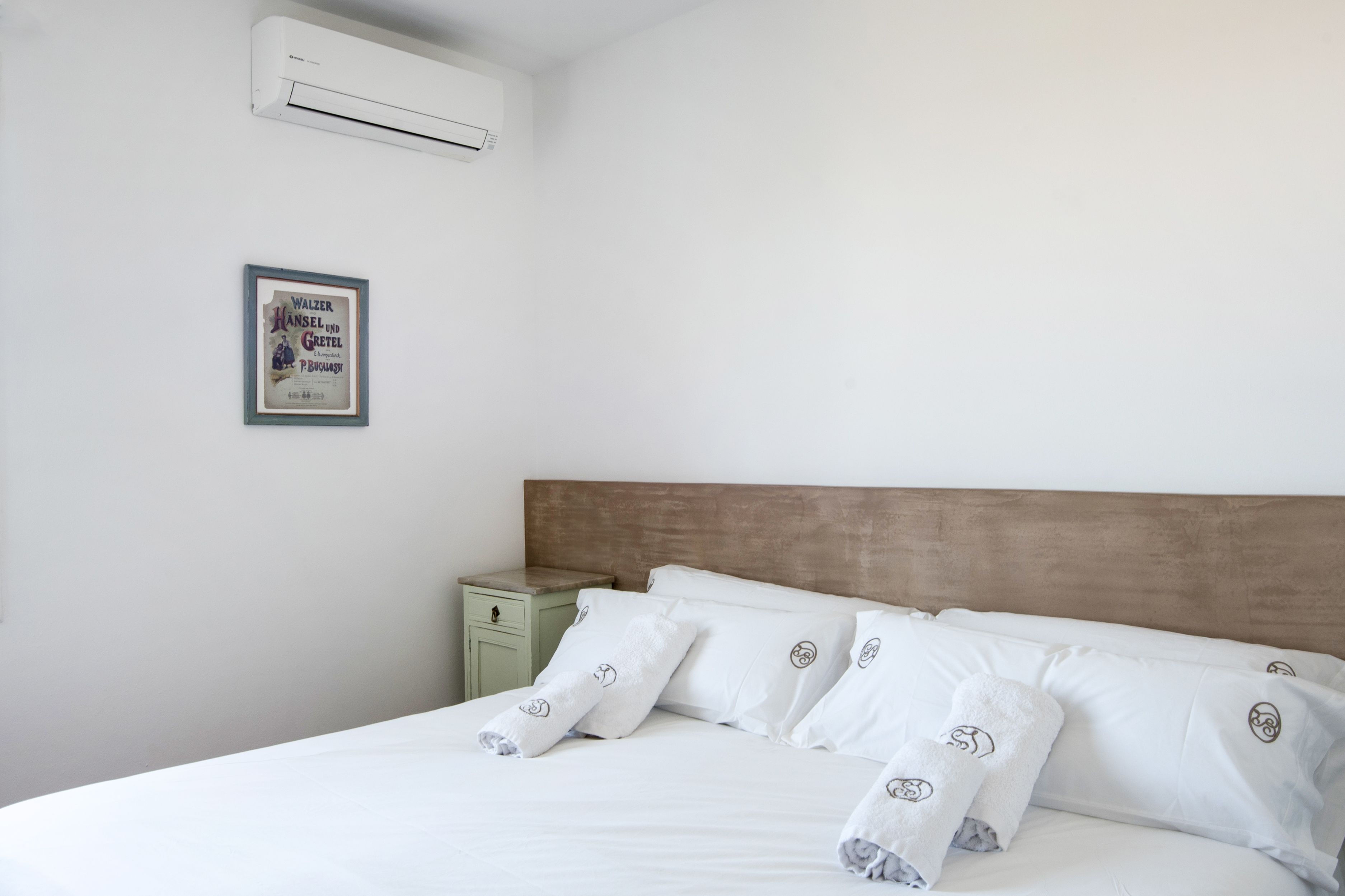 Una cama bien hecha con sábanas y almohadas blancas, una mesita de noche con lámpara, y un cuadro en la pared.