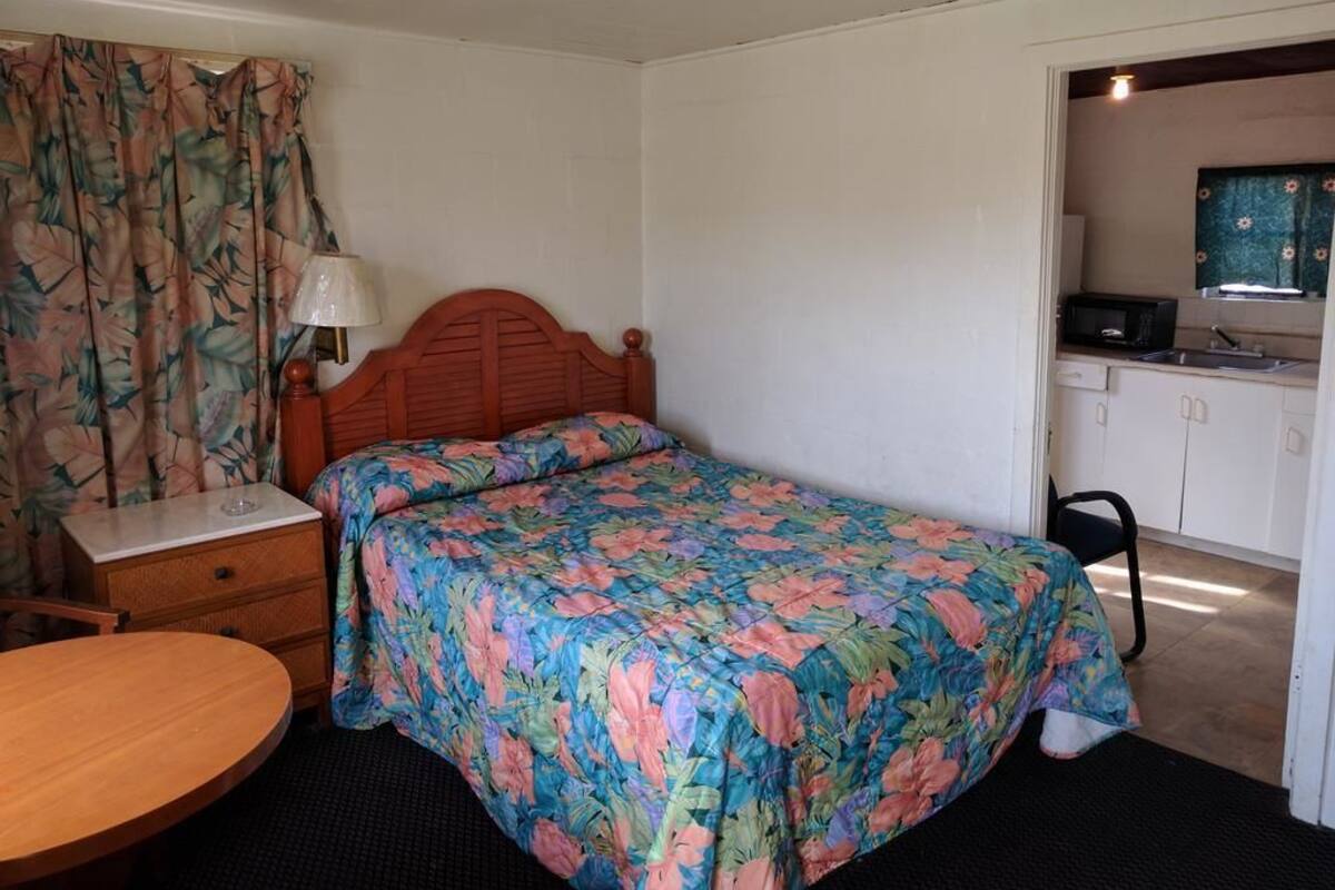A bedroom with a bed, a nightstand, a table, a chair, and a kitchenette in the adjacent room.