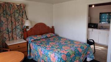 A bedroom with a bed, a nightstand, a table, a chair, and a kitchenette in the adjacent room.