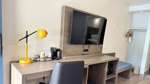Una habitación de hotel moderna con un escritorio de madera, una silla negra, un televisor de pantalla plana, una cafetera y un estante pequeño.