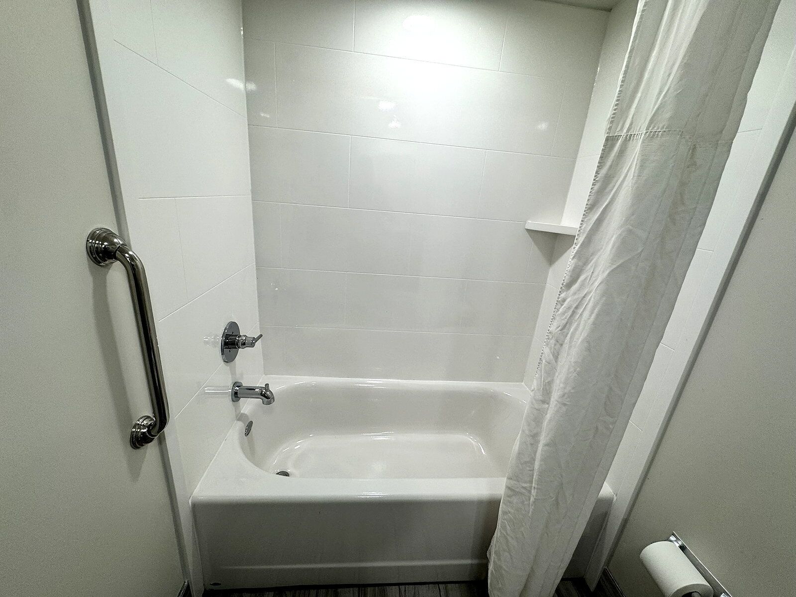 Une petite salle de bain aux murs recouverts de carreaux blancs, équipée d’une baignoire, d’un rideau de douche et d’un porte-rouleau de papier toilette.