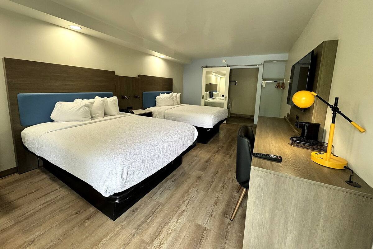 A hotel room with two beds, a desk, a chair, a lamp, and a TV.