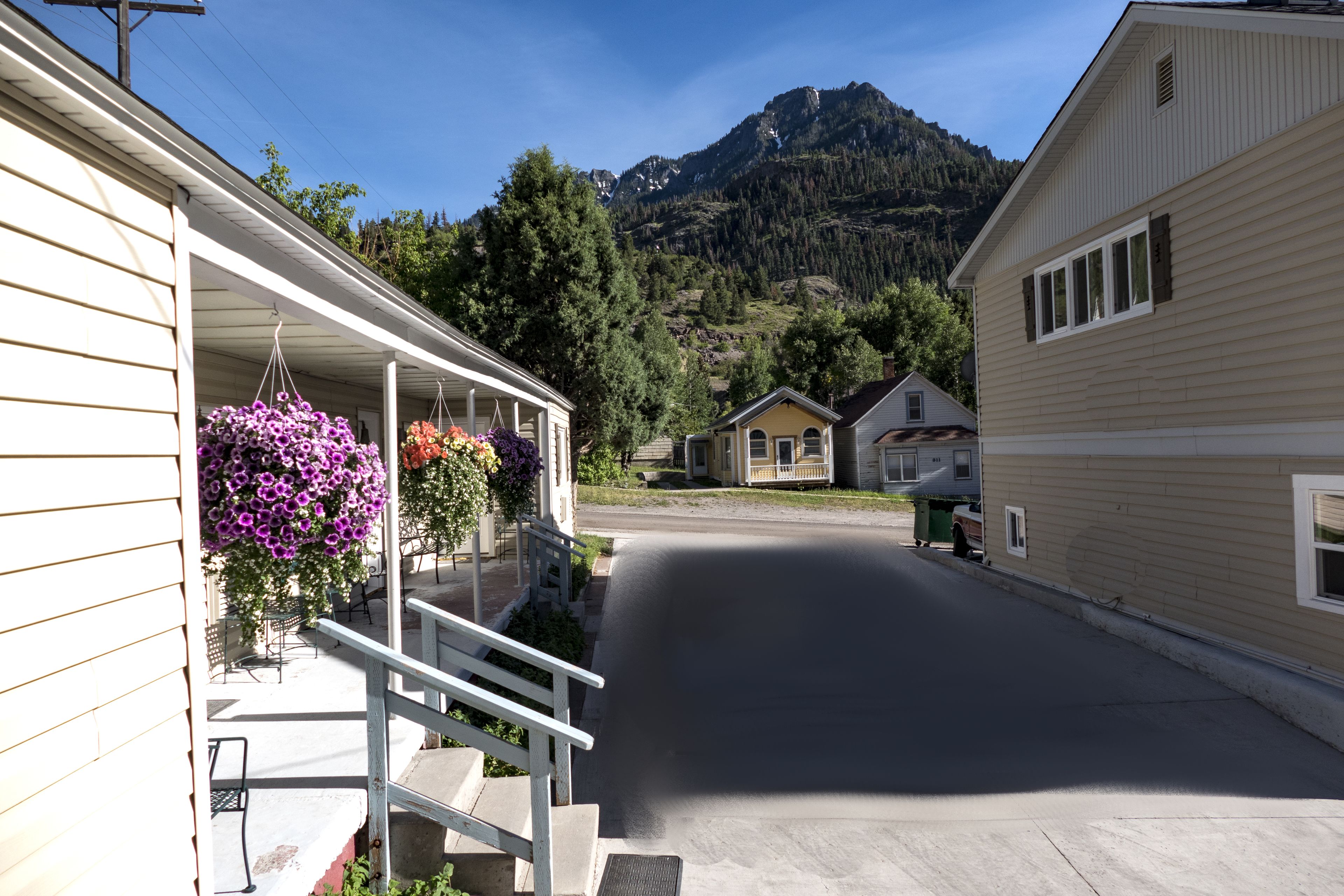 A quiet street with houses, a porch with hanging flowers, and a mountain in the background.