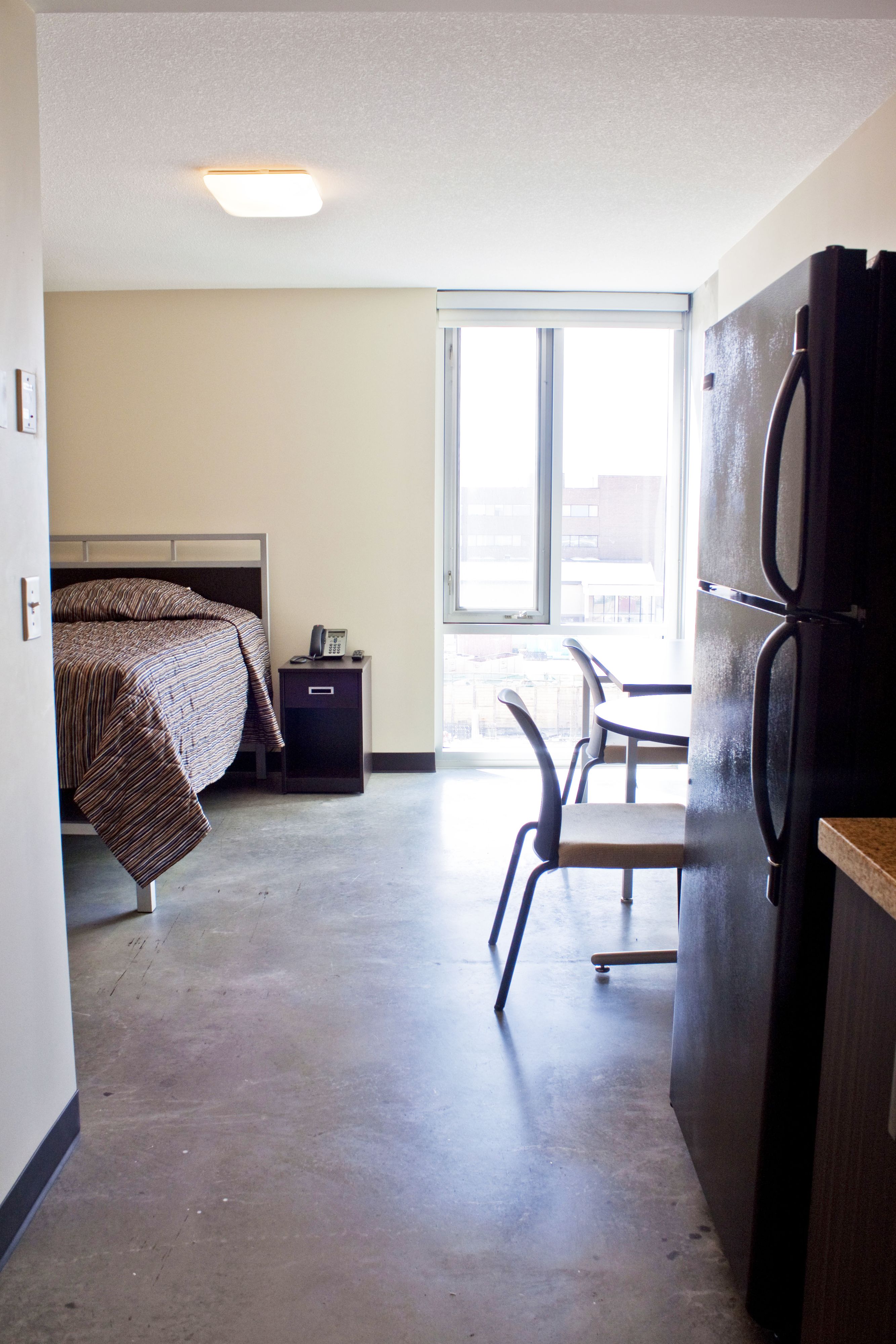 A hotel room with a bed, a desk, a chair, a refrigerator, and a window with a view of buildings.
