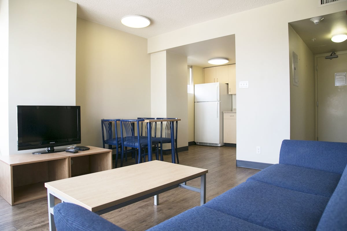 A modern living room with a flat-screen TV, a wooden coffee table, blue sofas, and a dining area with chairs and a table.