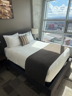 A hotel room with a large bed, a nightstand, a lamp, and a view of the city through a large window.