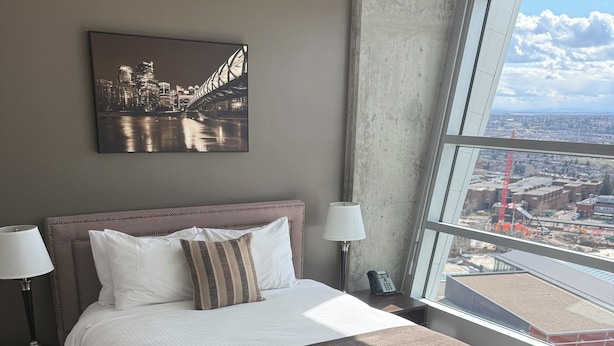 A modern hotel room with a large bed, two bedside lamps, a cityscape view through a large window, and a framed picture on the wall. - Residence & Conference Centre - Calgary (Calgary)