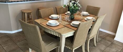 Un comedor con una mesa de madera dispuesta para seis personas, rodeado de sillas color beige. Se aprecia una cocina con armarios de madera y un microondas al fondo.