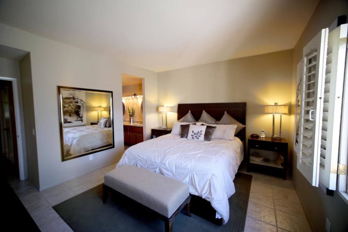 A hotel bedroom with a large bed, a bench, bedside tables, lamps, and a mirror.